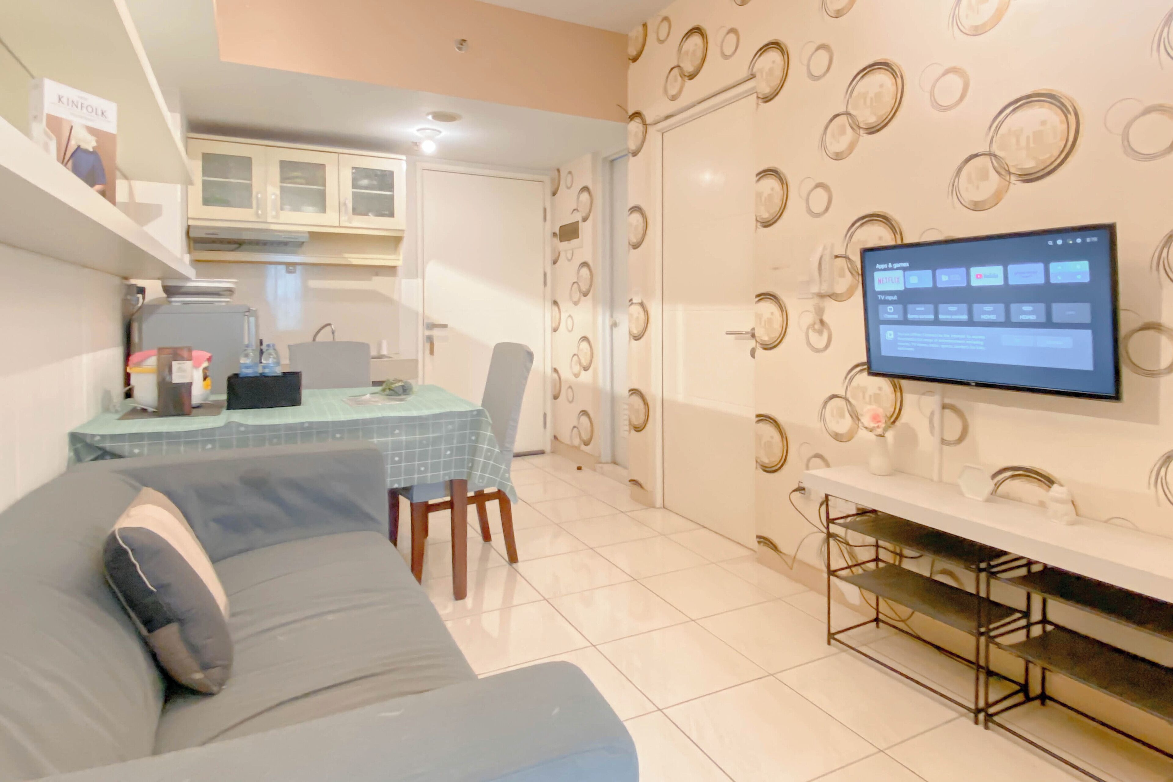 A modern living room with a sofa, a dining table with chairs, a TV mounted on the wall, and a kitchen area with cabinets and a refrigerator.