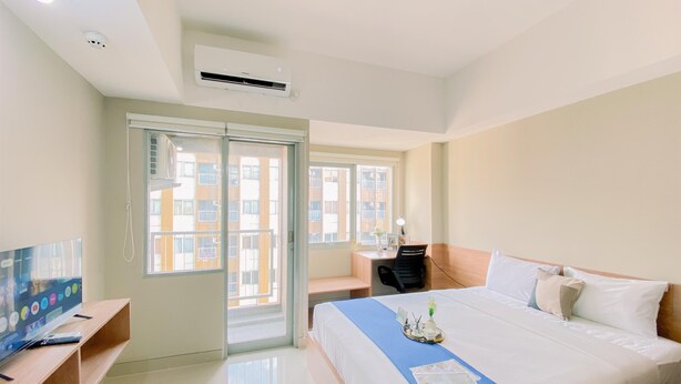 A hotel room with a bed, a desk, a television, and a window with a view of buildings. - Cozy Living Studio at LRT City Jatibening Apartment (Bekasi)