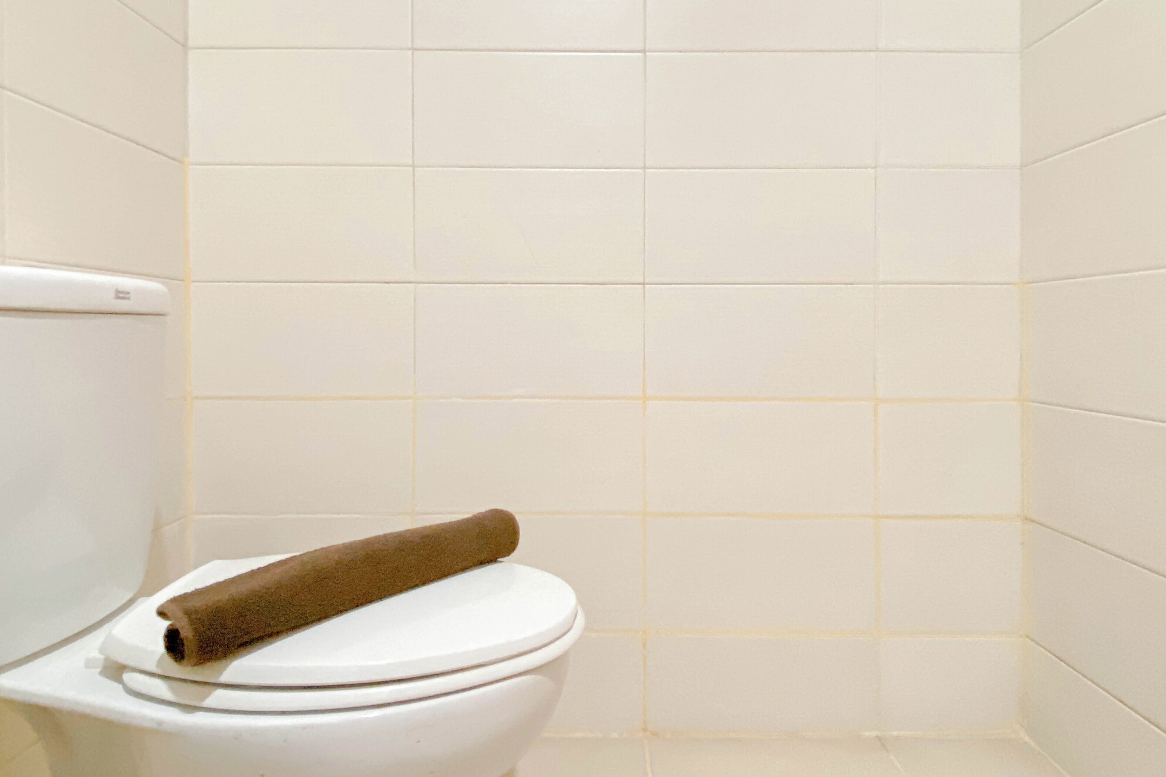 A toilet with a brown towel on the lid in a bathroom with white tiled walls.