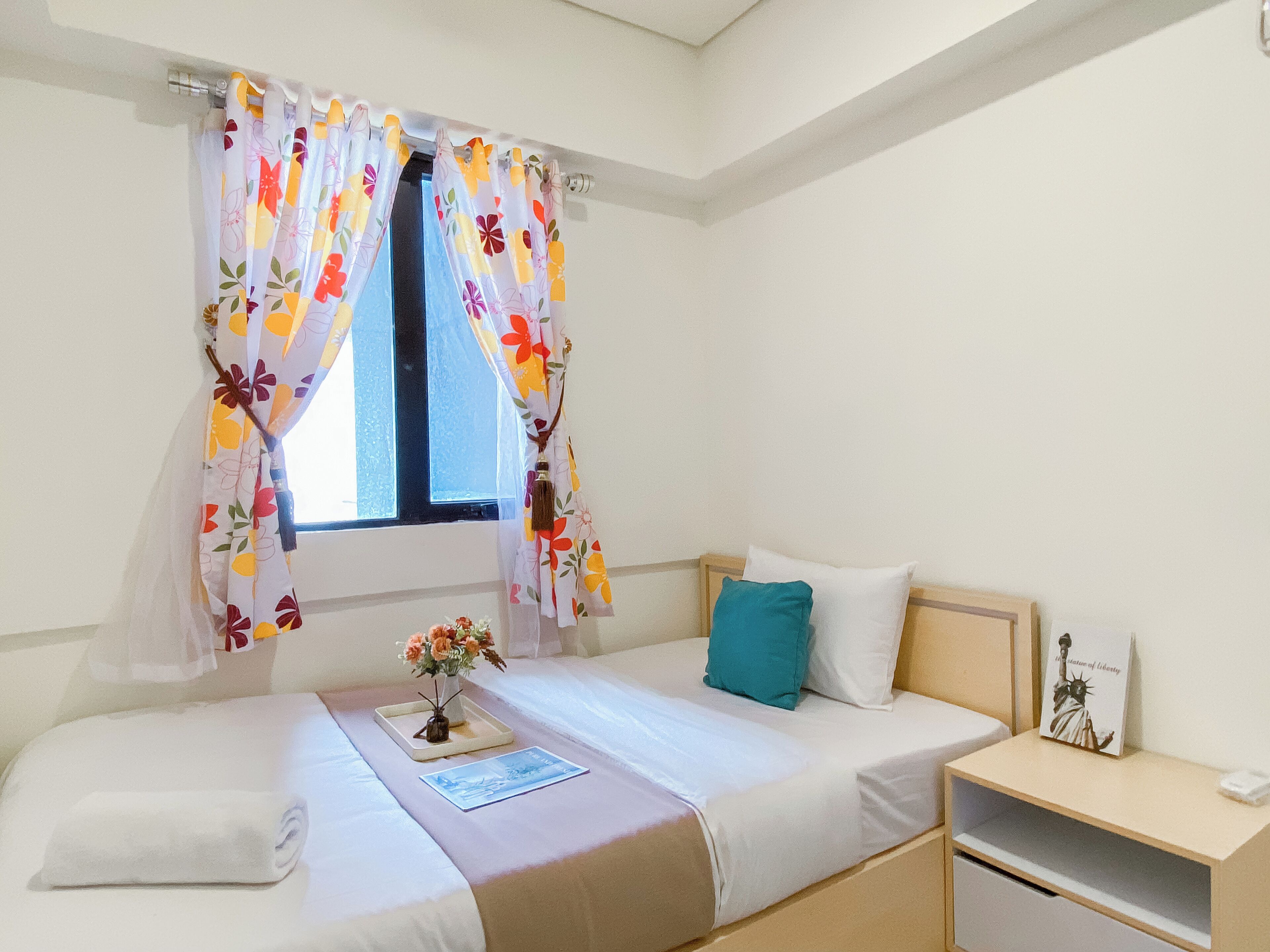 A bedroom with a bed, a bedside table, a nightstand, a window with floral curtains, and a small vase with flowers on the bed.