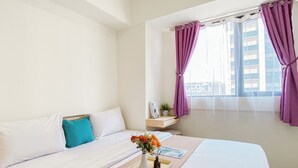 Quarto de hotel com uma cama, mesa de cabeceira com um vaso de flores, janela com cortinas roxas e vista para a cidade.