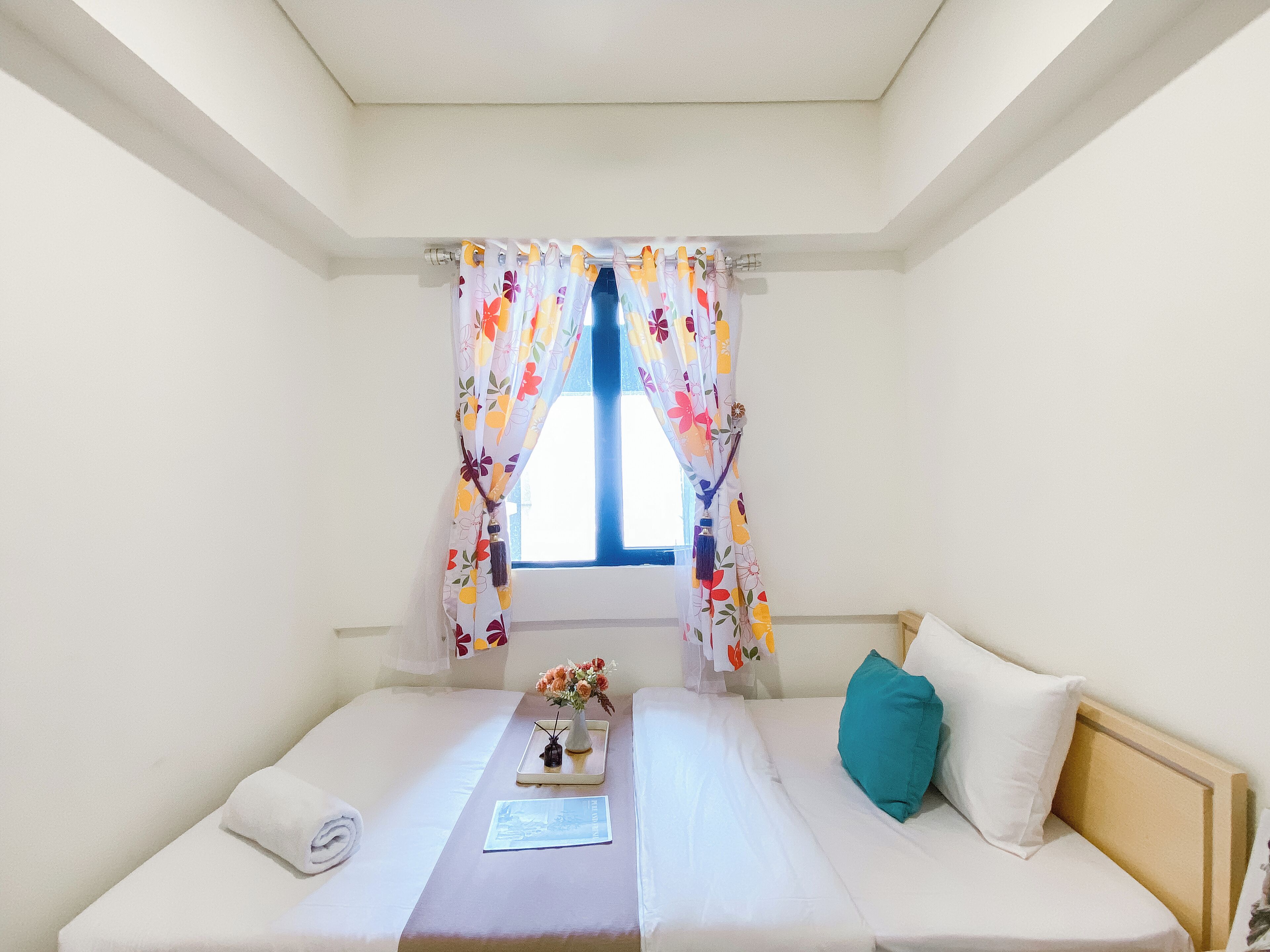 A single bed with a white headboard, a teal pillow, and a white towel. A small table with a vase of flowers and a tray is in front of the bed. The room has white walls and a window with floral curtains.