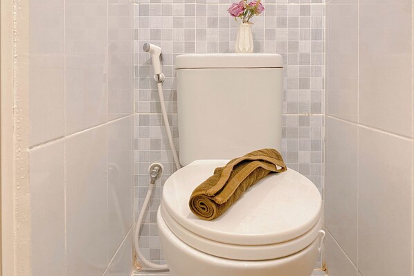 A toilet with a rolled-up towel on the seat, a shower hose, and a vase with flowers on the wall.