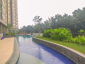 A swimming pool with a tiled edge, surrounded by greenery and a tall building in the background. - Good Deal and Comfy Studio at Transpark Cibubur Apartment (Depok)