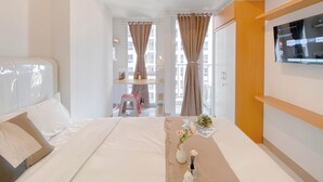 A hotel room with a bed, a TV mounted on the wall, a desk with a chair, and large windows with curtains. - Homey Studio Apartment at Tokyo Riverside PIK 2 (Teluknaga)