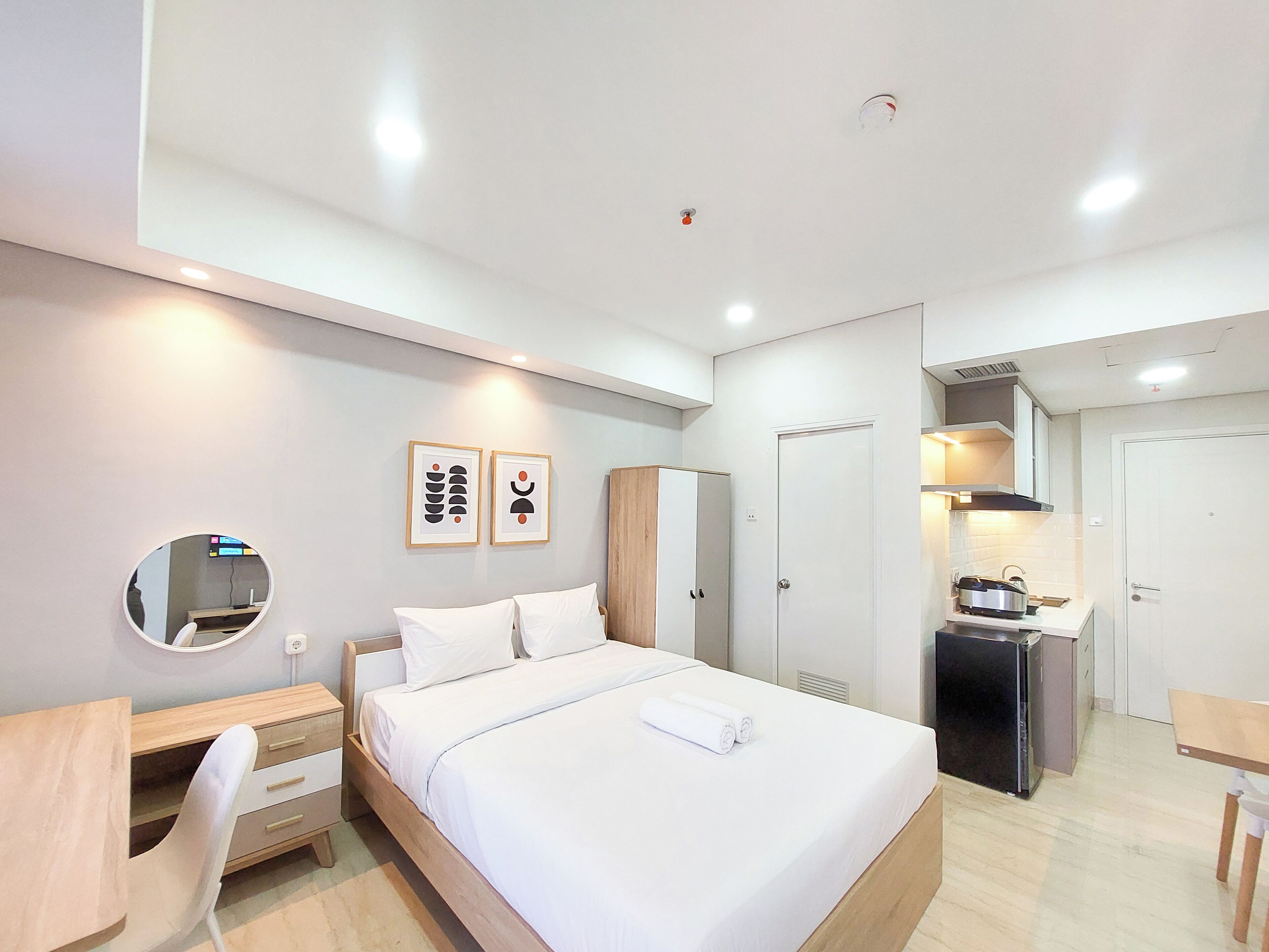 A modern hotel room with a bed, bedside table, chair, and a kitchenette area.