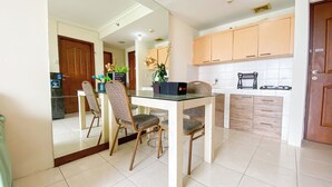 A dining area with a white table and patterned chairs, a kitchen with wooden cabinets, and a tiled floor. - Comfortable and Restful 2BR at Great Western Apartment (Tangerang)