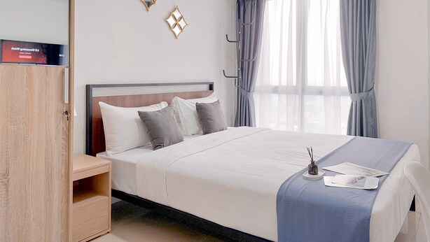 A neatly made bed with a blue bedspread, a wooden nightstand with a lamp, and a wardrobe. - Modern and Cozy Studio at Apartment Tamansari Bintaro Mansion (South Tangerang)