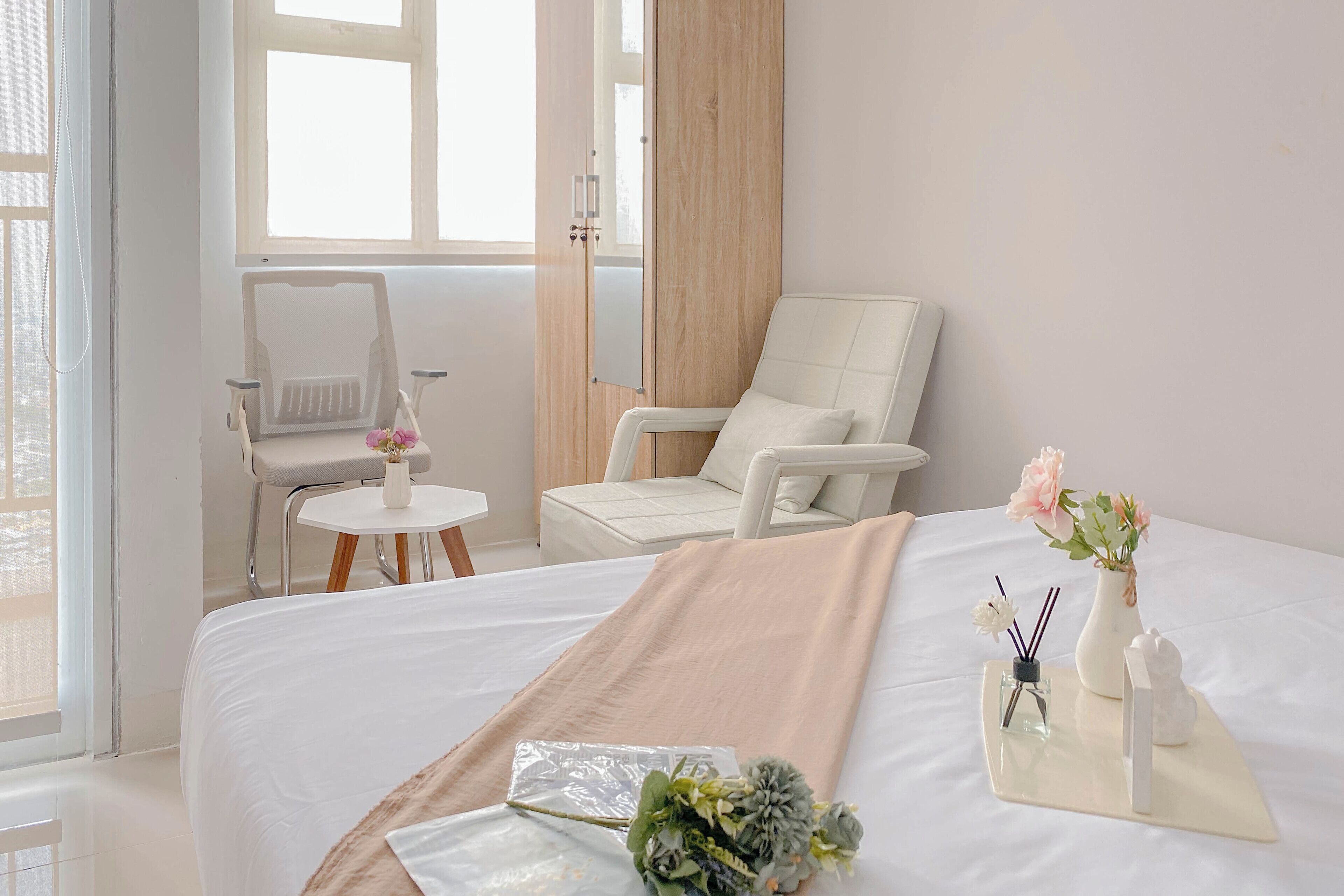 Une chambre d’hôtel avec un lit, un fauteuil blanc, une petite table sur laquelle est posé un vase de fleurs, et une fenêtre avec des rideaux.