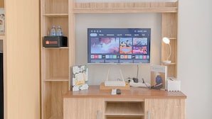 A wooden desk with a flat-screen TV displaying a streaming service interface, a remote control, a white desk lamp, and a small white speaker.