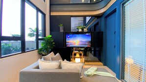 A modern living room with a grey sofa, a flat-screen TV, and a potted plant. - Elegant 2BR Loft Garden Villa at Collins Boulevard Apartment (Tangerang)