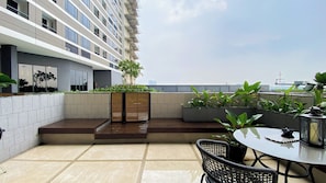 A rooftop terrace with a table and chairs, potted plants, and a view of the city skyline. - Elegant 2BR Loft Garden Villa at Collins Boulevard Apartment (Tangerang)