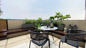 A rooftop terrace with a round table, four wicker chairs, potted plants, and a view of the city skyline. - Elegant 2BR Loft Garden Villa at Collins Boulevard Apartment (Tangerang)