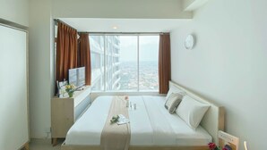 A hotel room with a large bed, a desk with a TV, and a view of the city through the window. - Cozy Studio at Grand Kamala Lagoon Apartment near Mall (Bekasi)