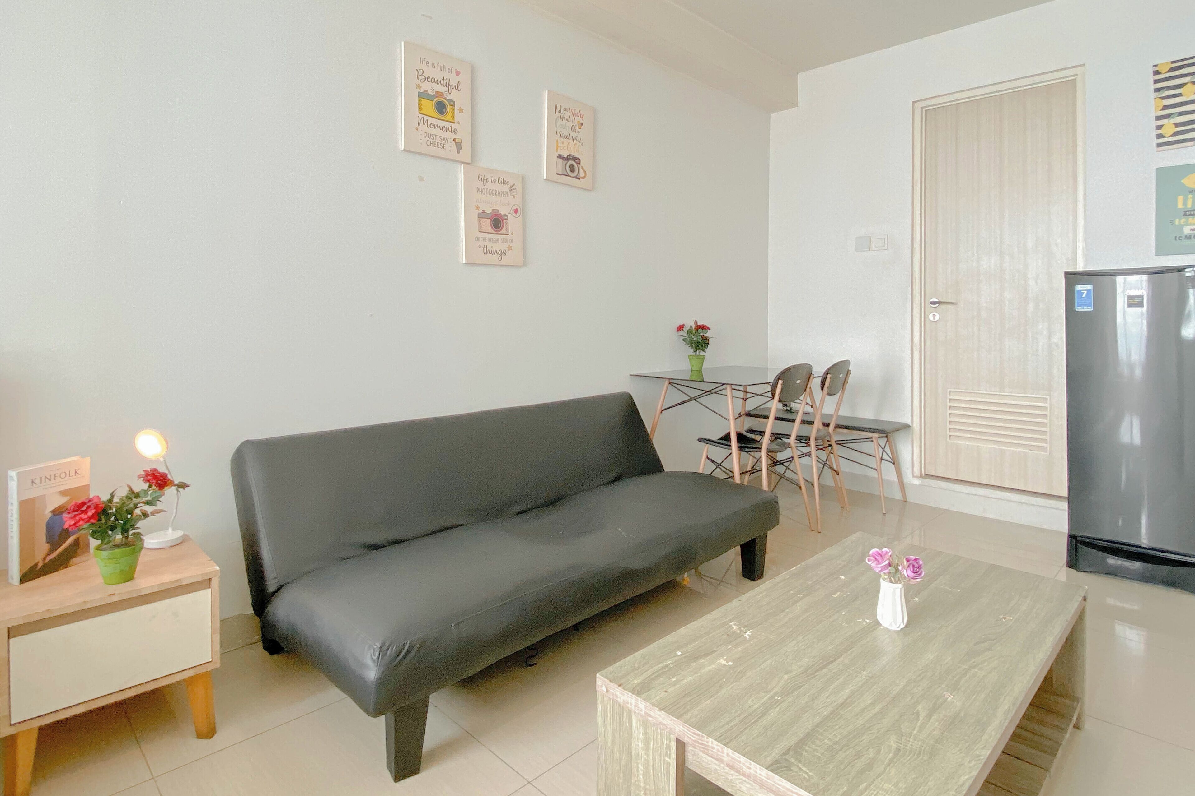 A modern living room with a grey sofa, a wooden coffee table, and a small side table with a lamp and a plant. There is a dining area with a table and chairs, and a refrigerator in the corner.