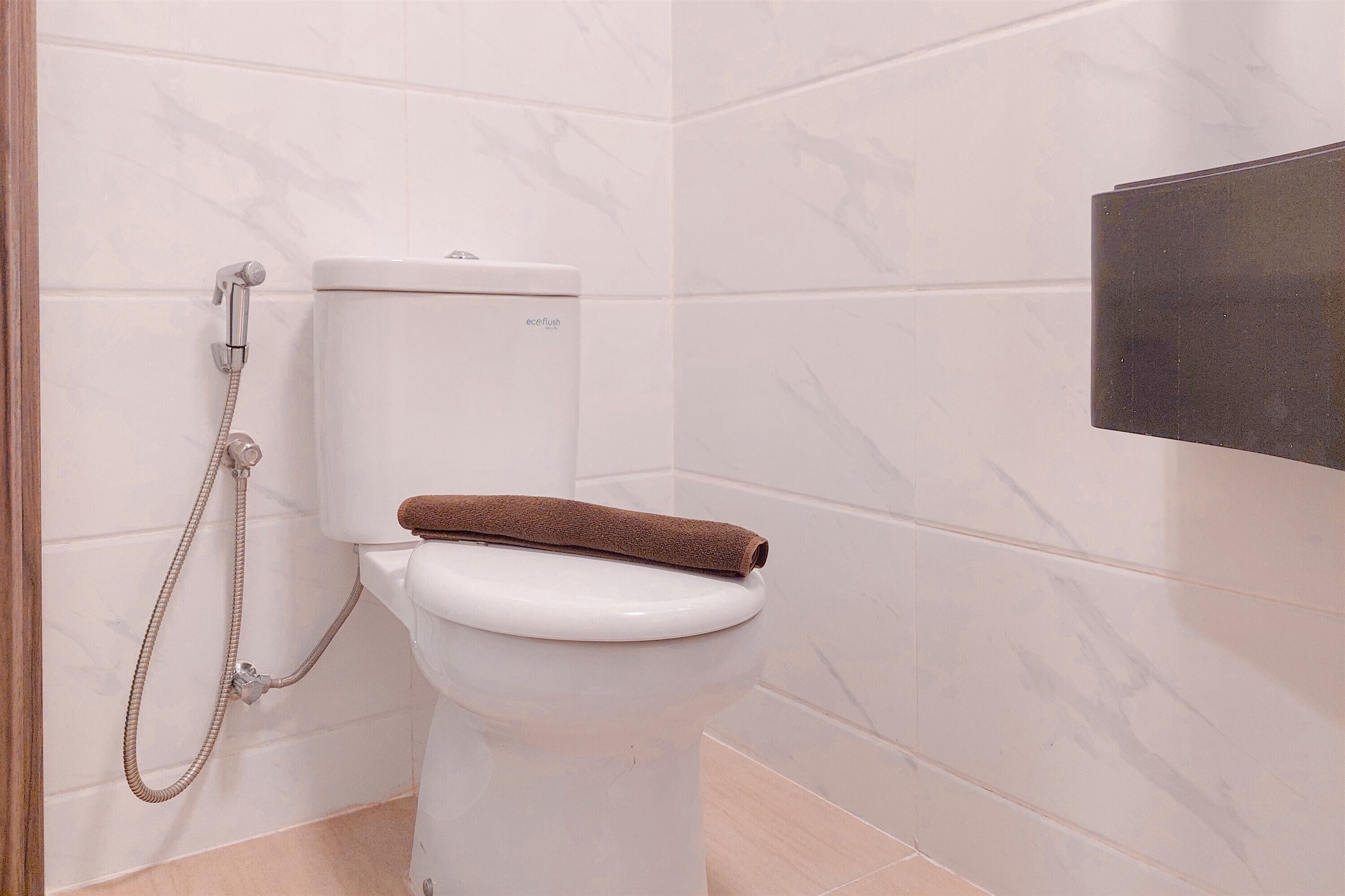 Eine weiße Toilette mit einem braunen Handtuch darauf, eine Handbrause und eine schwarze Platte an der Wand.
