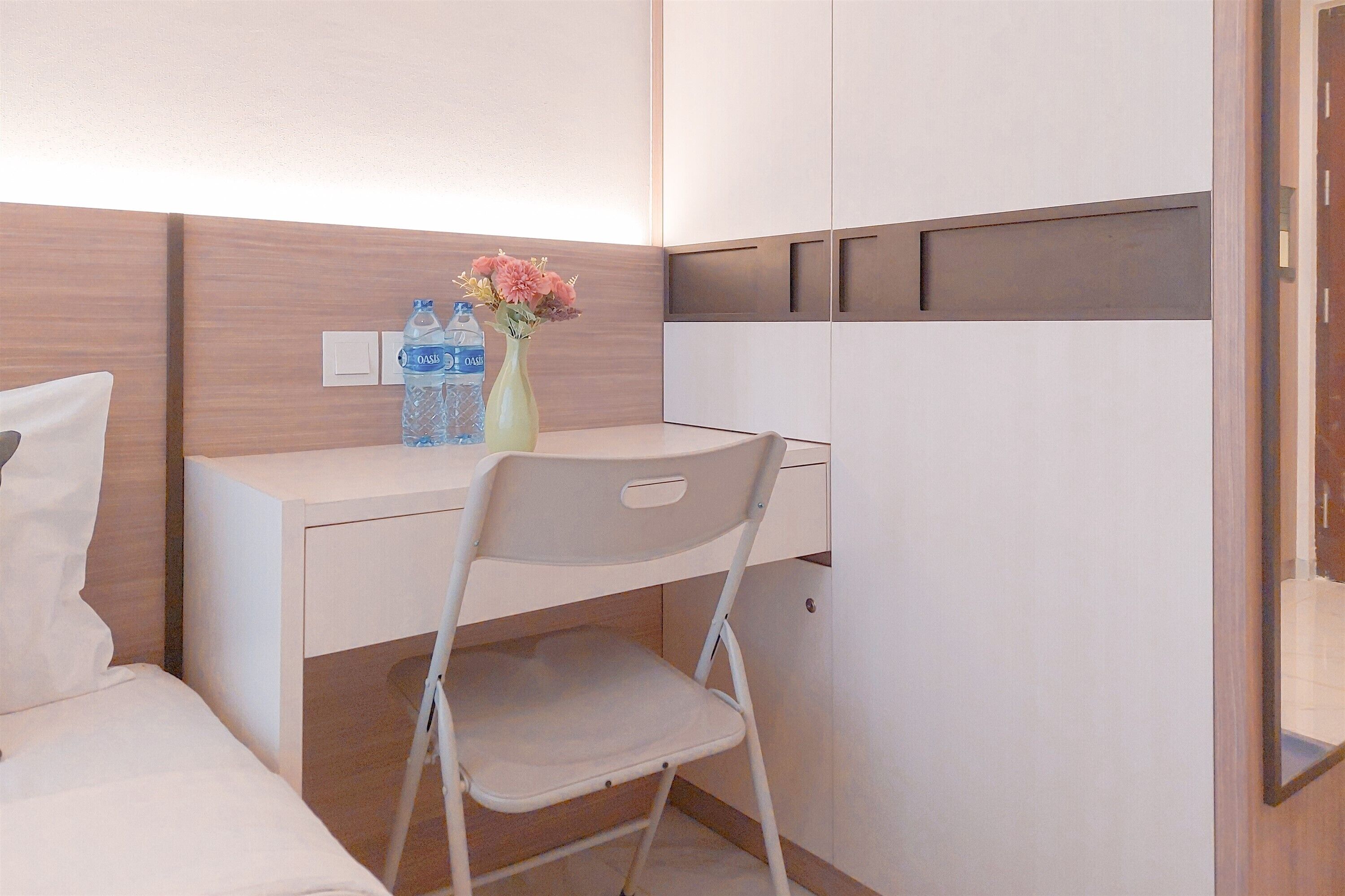 Une chambre d’hôtel de taille réduite, comprenant un lit, un bureau, une chaise, un vase avec des fleurs et une bouteille d’eau.