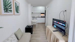 Un espacio habitable pequeño con un sofá, un televisor sobre una mesa y una kitchenette al fondo.