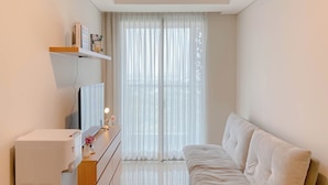 A bedroom with a bed, a dresser, a shelf with books and a lamp, and a window with sheer curtains.