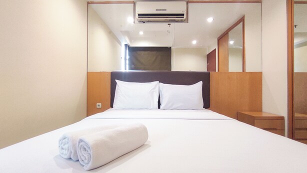 A hotel room with a bed, white linens, a rolled towel, an air conditioning unit, and a mirror. - Good Place and Tidy 2BR at High Point Serviced Apartment (Surabaya)