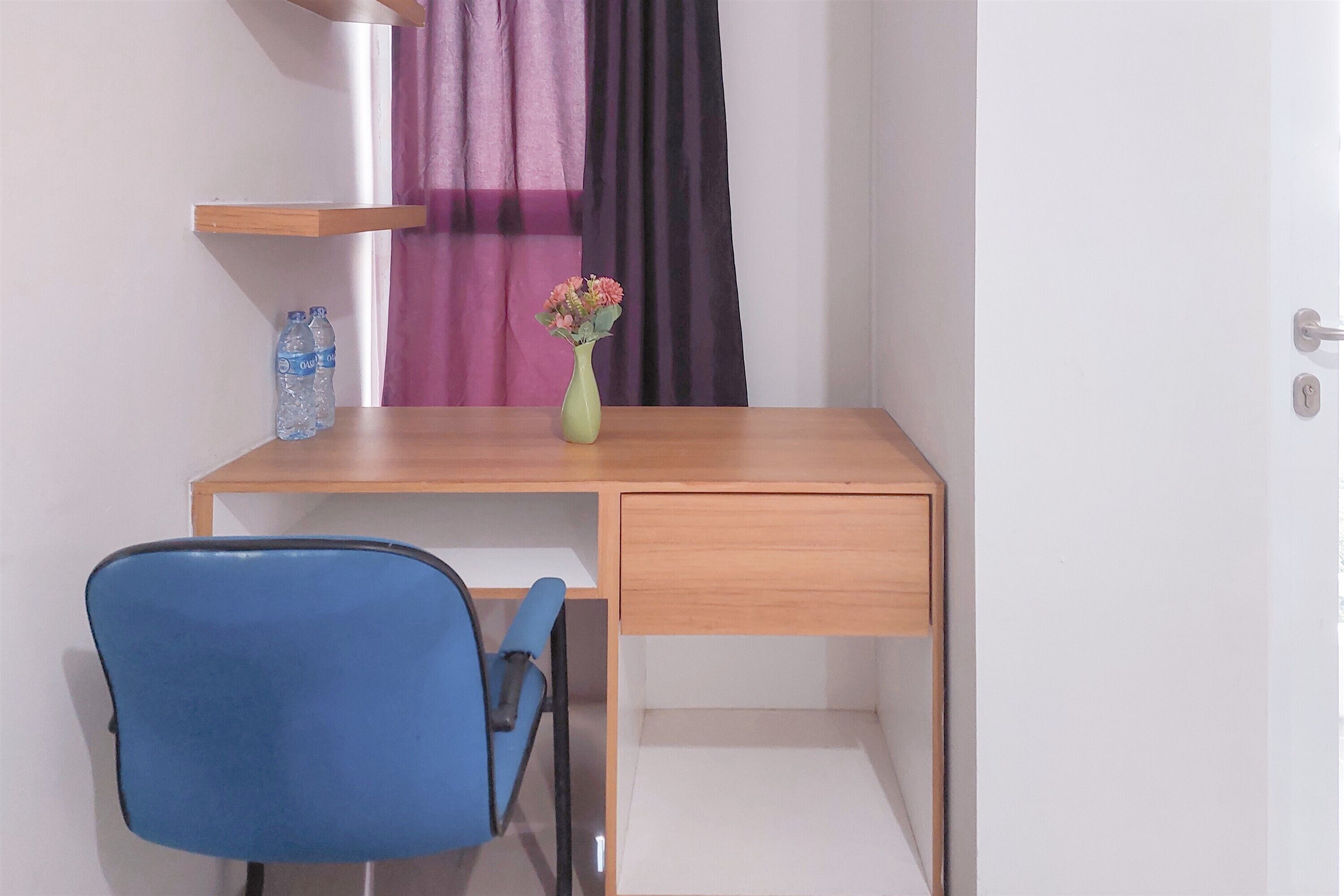 Een kleine, eenvoudige kamer met een houten bureau, een blauwe stoel, een vaas met bloemen en een waterfles op een plank.