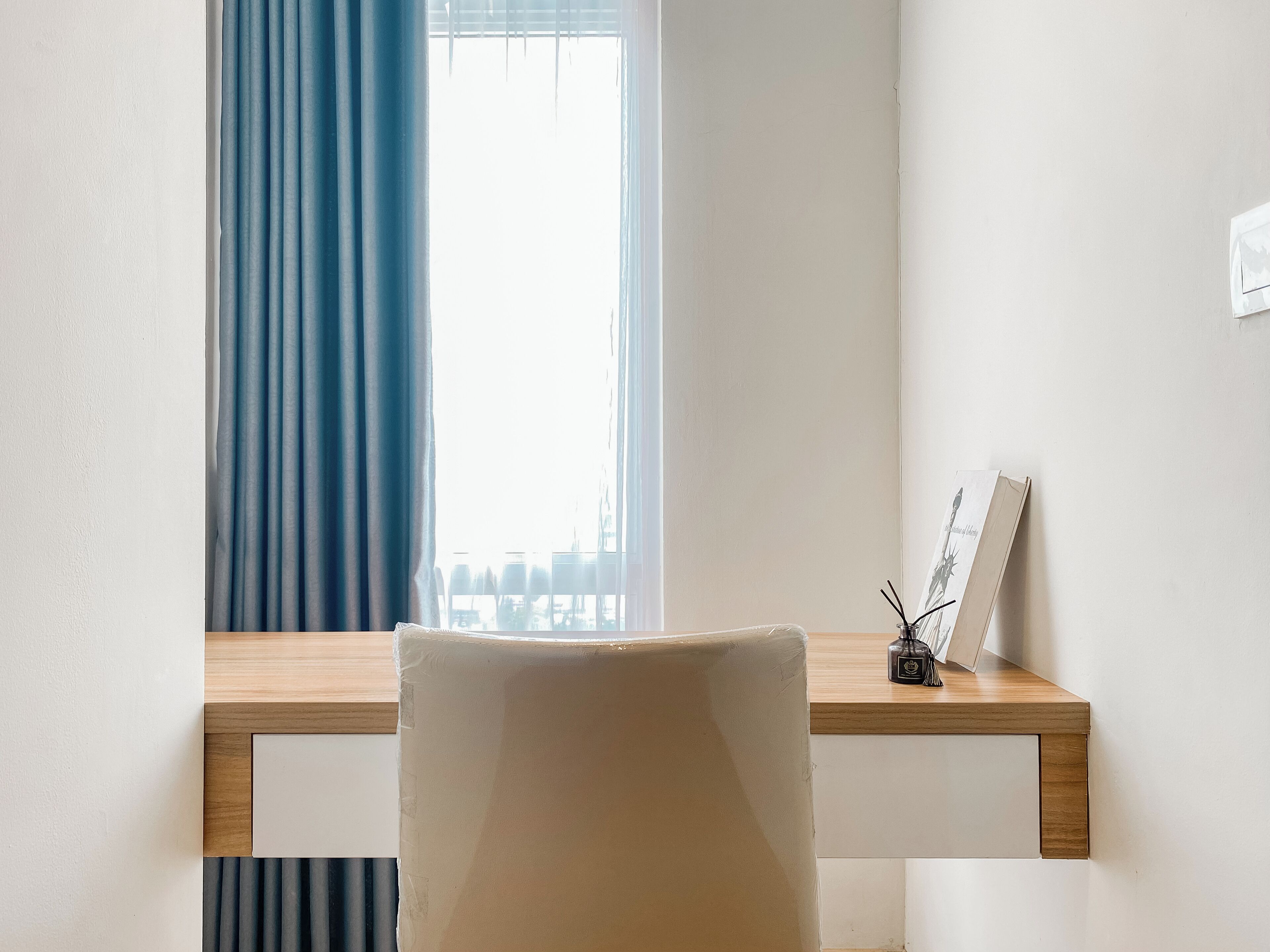 A room with a wooden desk, a chair, a window with blue curtains, and a picture frame with a candle holder on the desk.