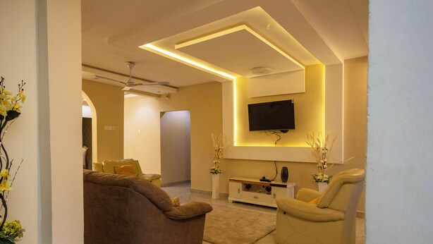 Een moderne woonkamer met een bank, een verstelbare fauteuil, een flatscreen-televisie aan de muur en een plafondlamp.