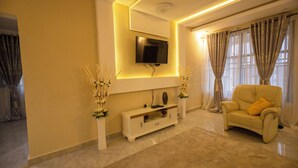 Een woonkamer met een flatscreen televisie aan de muur, een beige bank met een geel kussen, en een witte console tafel met decoratieve planten.