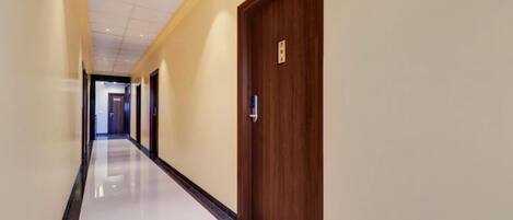 A long, well-lit hallway with a polished floor, leading to a wooden door with a handle and a small sign above it.