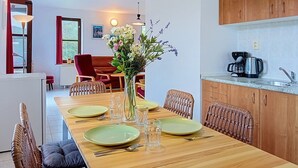 Un coin repas avec une table en bois dressée pour quatre personnes, un vase rempli de fleurs et une cuisine dotée de meubles en bois et d’une machine à café.