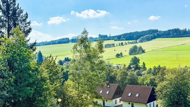 Un paysage rural avec une maison blanche dotée d’un toit en tuiles, entourée d’une végétation luxuriante et de champs.