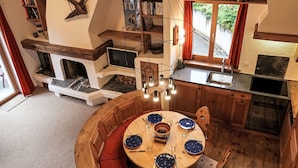 A cozy, rustic interior with a wooden dining table set for four, a fireplace, and a kitchen area with a window overlooking greenery.