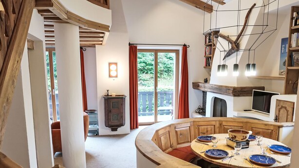 A cozy, rustic dining area with a wooden table set for four, surrounded by wooden chairs. A built-in fireplace and a bookshelf are visible in the background.