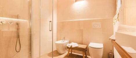 A modern bathroom with a glass shower enclosure, a bidet, a toilet, and a wooden stool.