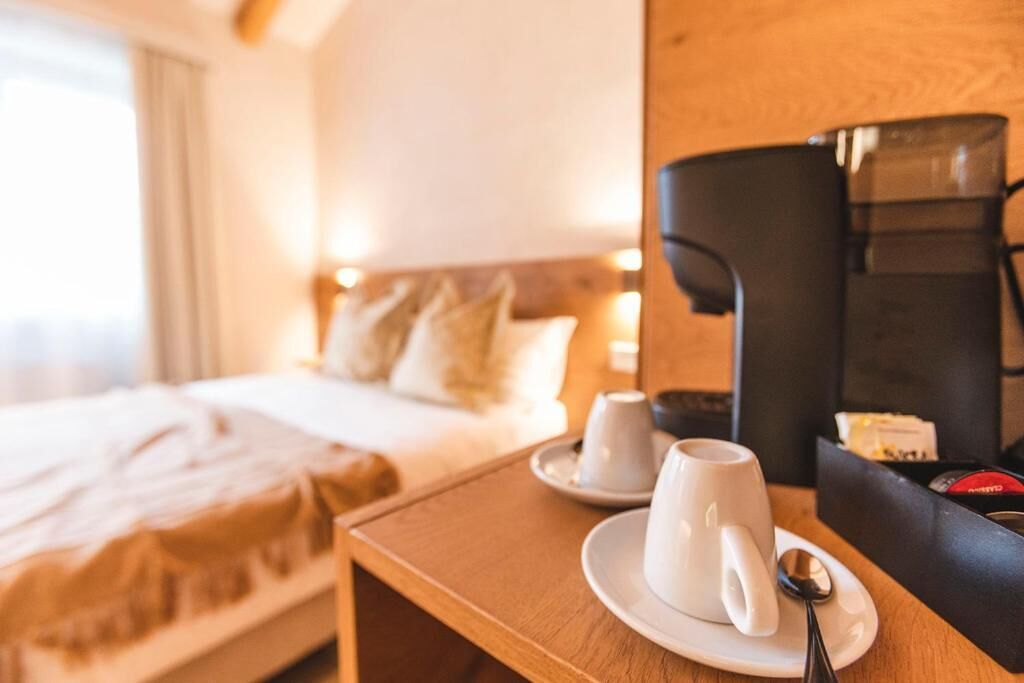 A hotel room with a bed, a bedside table with a coffee maker, cups, and a spoon.
