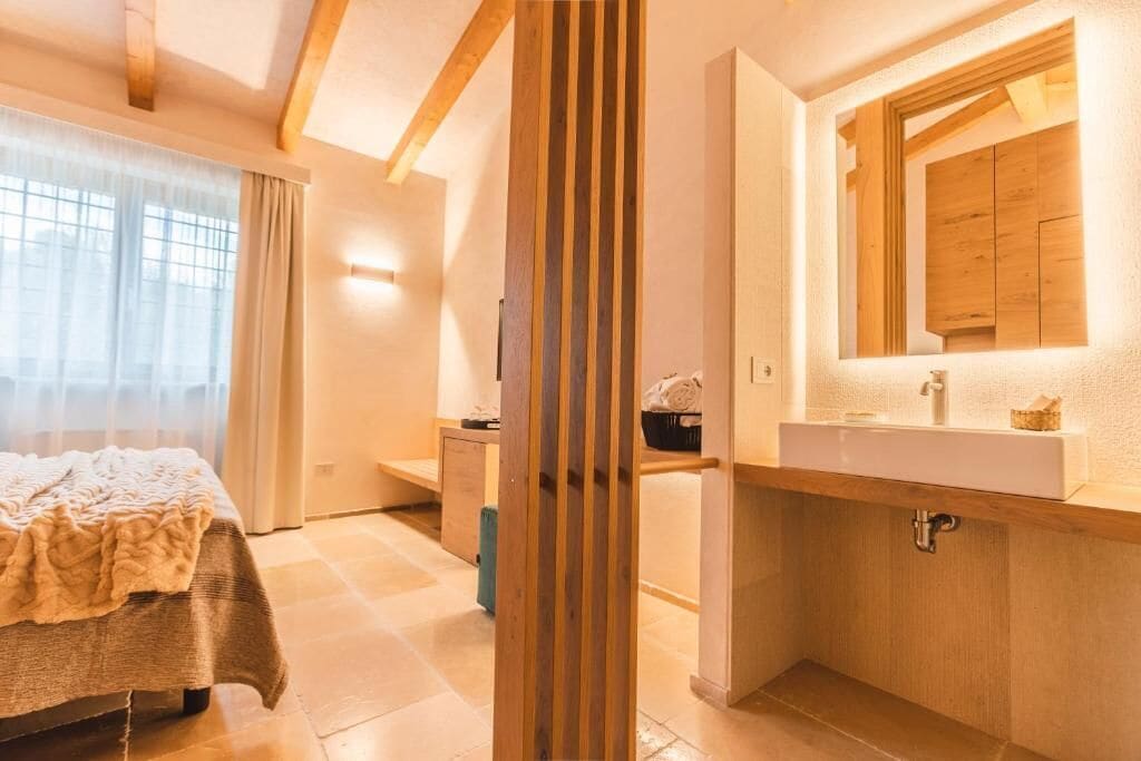 Ein modernes Schlafzimmer mit Bett, Nachttisch, einer Holzsäule, einem Badezimmer mit Waschbecken und Spiegel sowie einem Fenster mit Vorhängen.