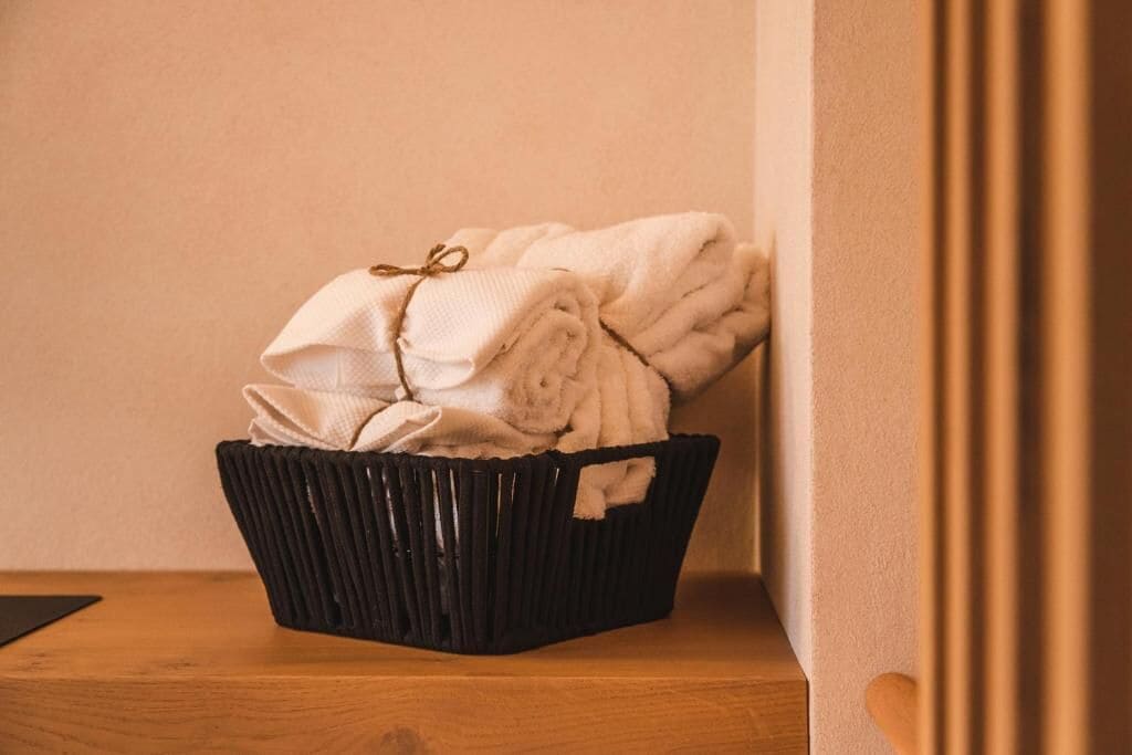 Una cesta negra llena de toallas dobladas sobre una superficie de madera, con una pared de fondo.