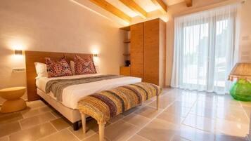 Un dormitorio con cama, mesitas de noche, un banco, vigas de madera, una ventana con cortinas y una lámpara.