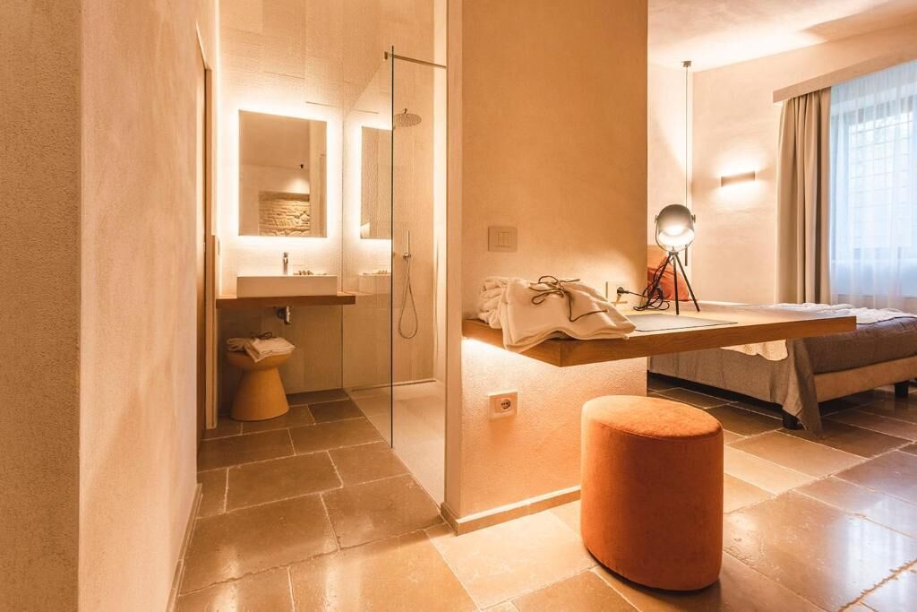 A modern bathroom with a glass shower enclosure, a vanity with a mirror, and a bedside table with a lamp.