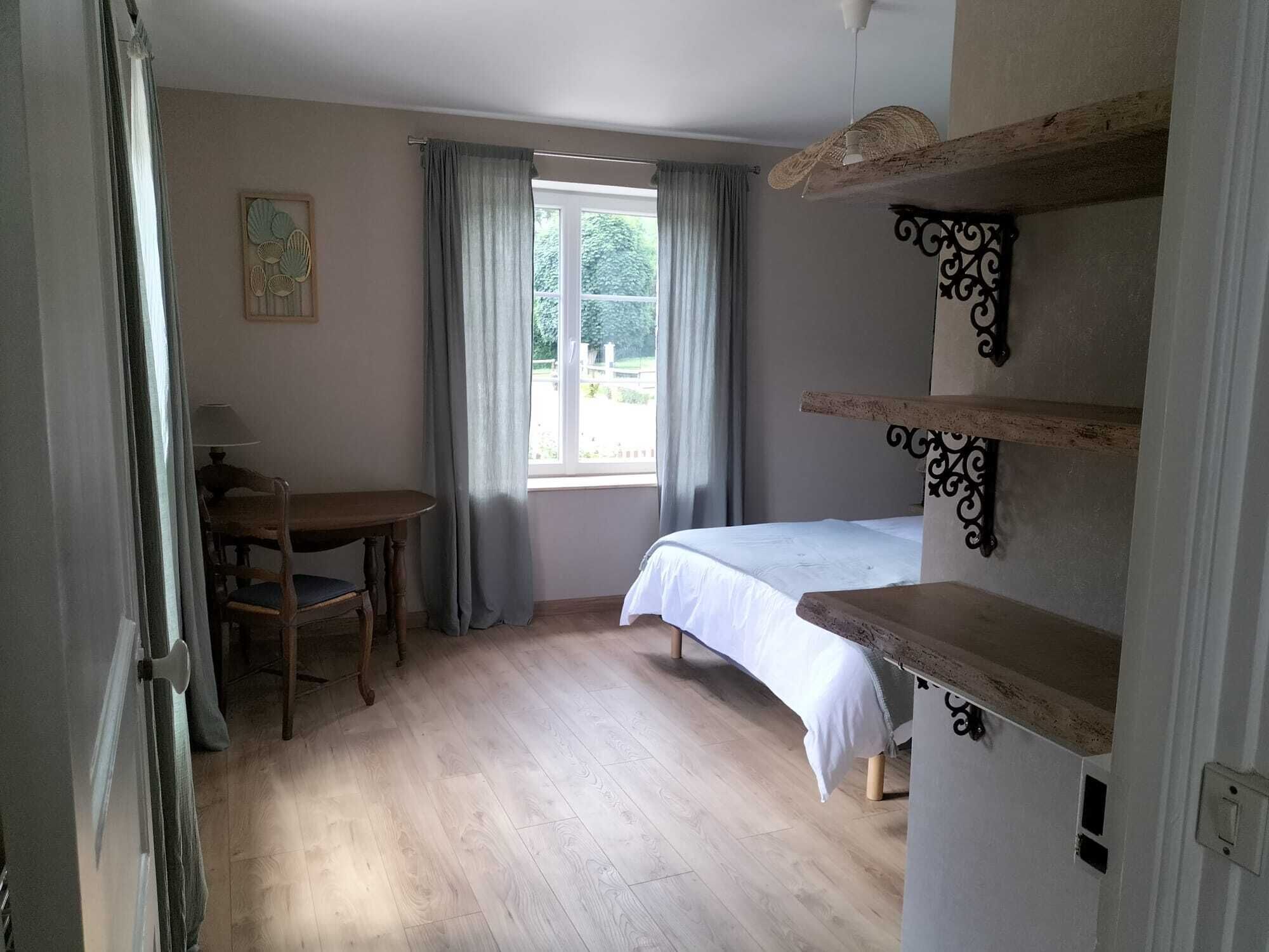 A bedroom with a bed, a desk, and a chair. There is a window with curtains, a wall-mounted shelf, and a door.