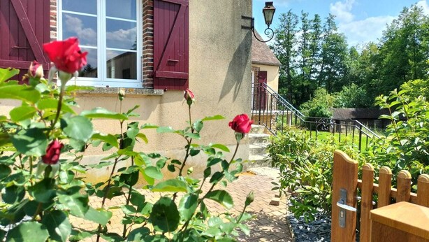 Ein Haus mit Ziegel Fassade, ein Fenster mit roten Lamellen, ein Holztor und ein Rosengarten.