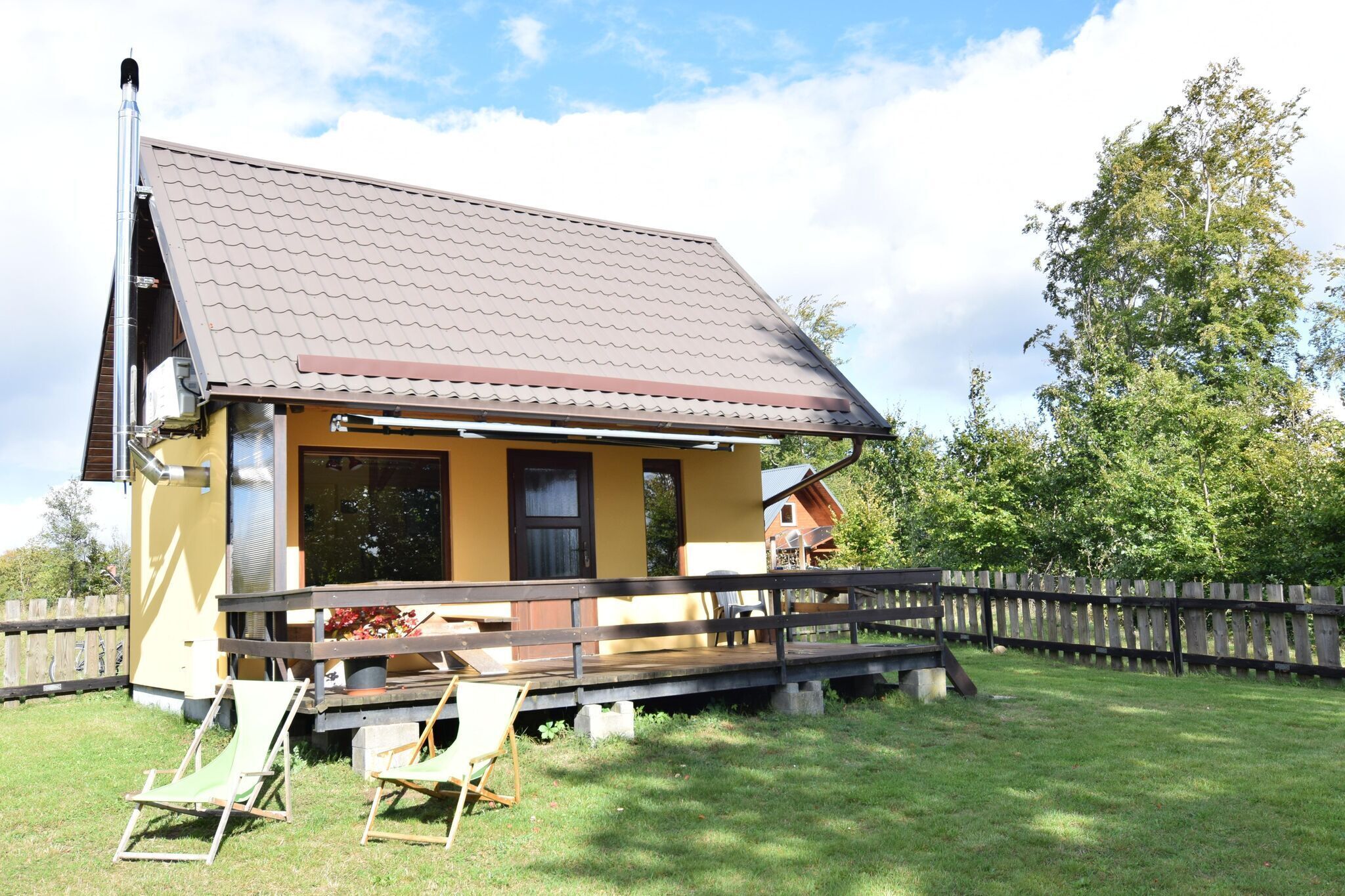 Une maison jaune avec un toit marron, une terrasse en bois et une clôture en bois.