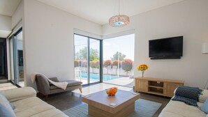 A modern living room with a sofa, a coffee table with oranges, a TV, and a view of a swimming pool. - Casa Madeira - Stunning Villa in Carvoeiro (Carvoeiro)
