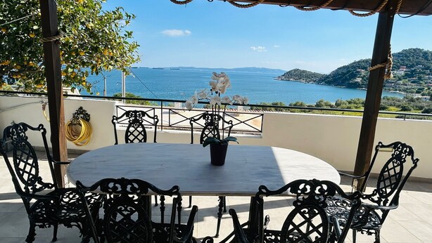 Un espace extérieur aménagé, abrité et doté de tables et de chaises, offrant une vue sur un paysage côtier avec la mer et des collines lointaines.