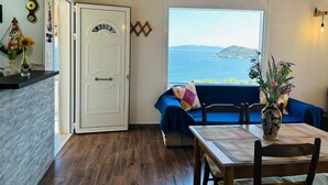 Une pièce avec un plafond en bois, une grande fenêtre donnant sur la mer, une table à manger avec des chaises et un canapé bleu.