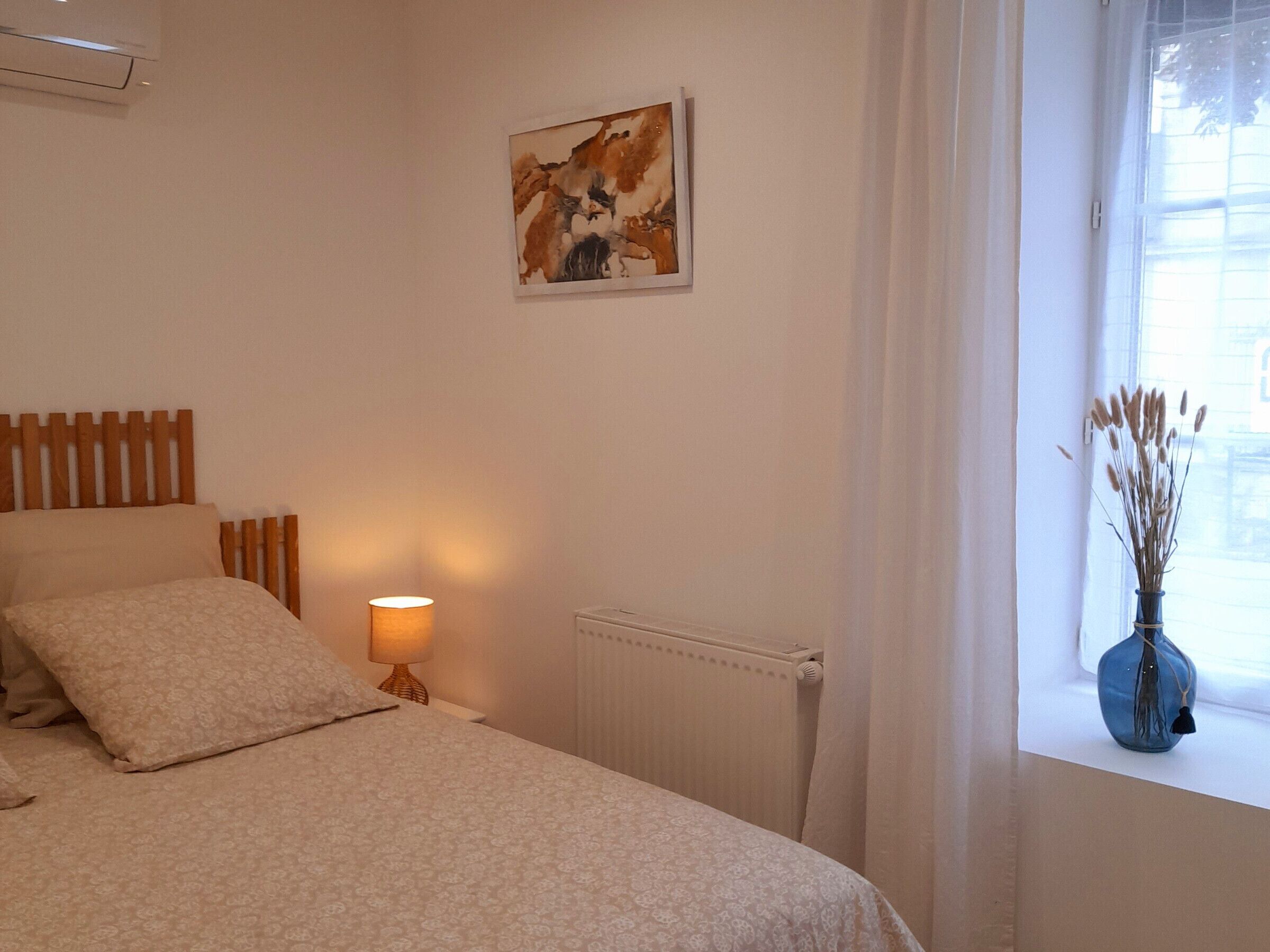 Un dormitorio con cama, lámpara de noche, radiador, ventana con cortinas y un cuadro en la pared.