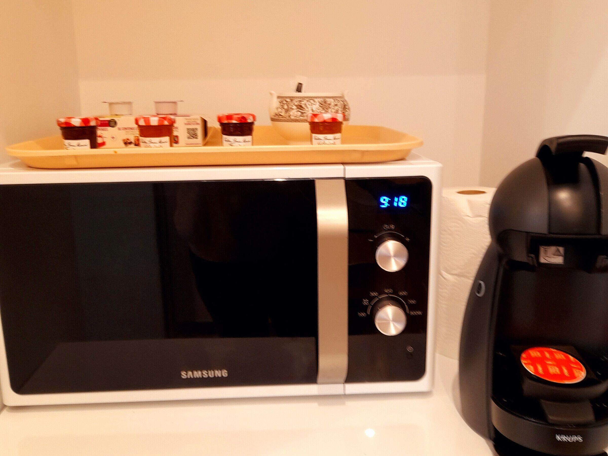 A Samsung microwave with a digital clock displaying 9:18, a coffee maker, and a tray with jars of condiments.