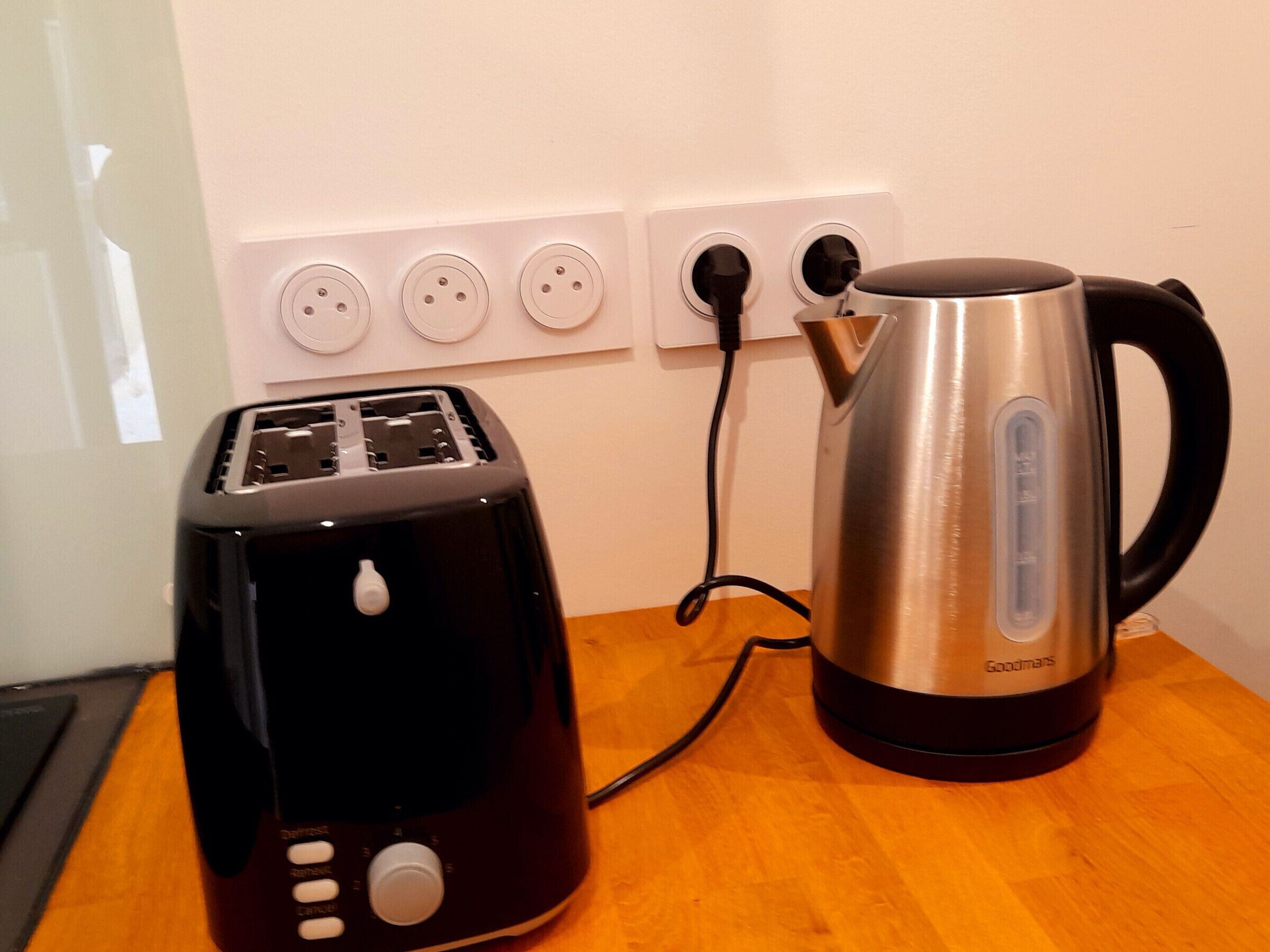 A black toaster and a stainless steel kettle on a wooden table, connected to a power outlet.