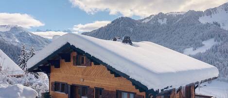 A wooden chalet with a snow-covered roof, set against a mountainous backdrop.