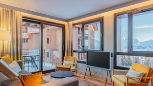 Een moderne woonkamer met een bank, stoelen, een televisie en grote ramen met uitzicht op een gebouw en bergen.
